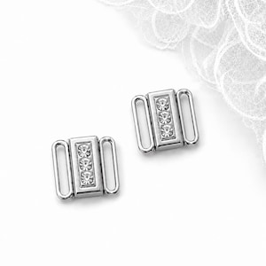 May include: Two rectangular silver-toned metal clothing fasteners, each featuring a central vertical row of three clear, round, faceted rhinestones and two side loops for attachment to fabric or straps, presented on a plain white background.