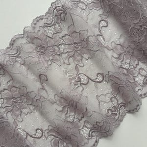 Könnte beinhalten: Nahaufnahme eines Stücks grauen Spitzenstoffs. Die Spitze weist ein Blumenmuster mit detaillierten, strukturierten Designs auf. Die Kanten sind gewellt und der Stoff wirkt zart und halbtransparent, geeignet für Kleidung oder Bastelprojekte.