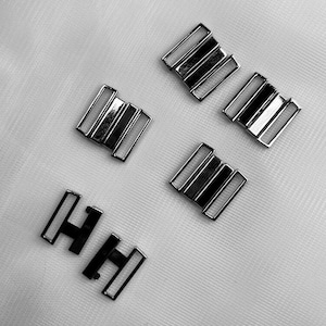 May include: A collection of silver-colored metal bra strap adjusters and hooks. The adjusters have a rectangular shape with three parallel bars. The hooks are shaped like the letter "H". The items are arranged on a white, textured surface.