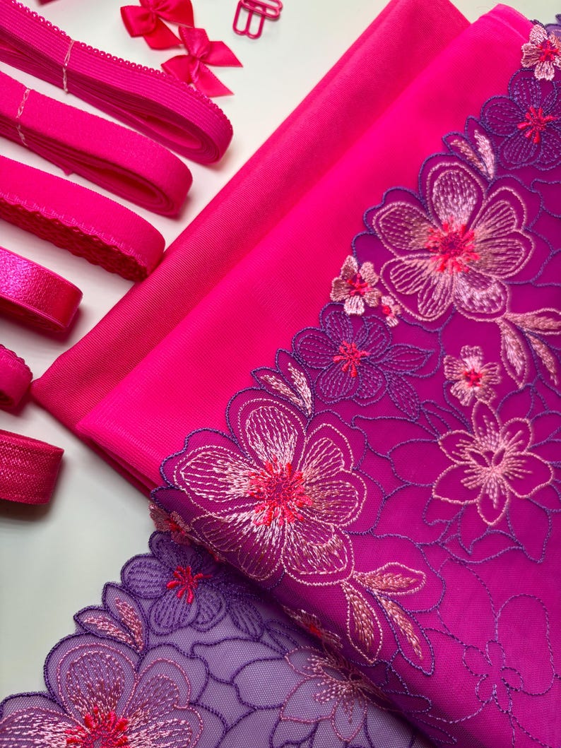 Op de afbeelding: Roze en paarse stof met bloemenborduursel. De stof is gelaagd met een roze mesh stof bovenop een effen roze stof. Er is ook een roze elastiek en een roze lint met een strik.