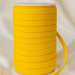 May include: A spool of bright yellow elastic ribbon. The ribbon is tightly wound and sits on a white base. The word "YELLOW" is printed in black at the top of the image. The ribbon is unwound slightly, showing its flexibility.