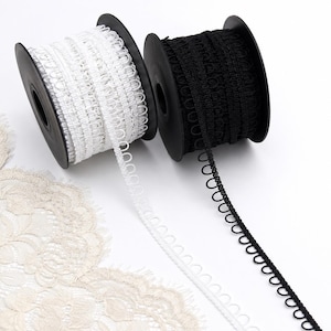 May include: Two black plastic spools holding button loop tape, one with white trim and one with black trim. The tape features a continuous row of small, uniform fabric loops designed for fastening, extending from each spool onto the surface.