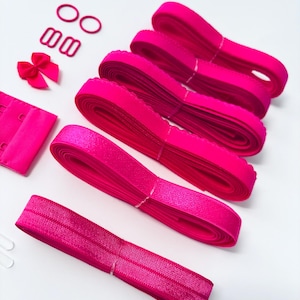 Op de afbeelding: Een set roze bh-bandverlengstukken met een roze strik, twee kleine roze ringen en twee grotere roze gespen. De verlengstukken zijn gemaakt van elastiek en hebben een gegolfde rand.
