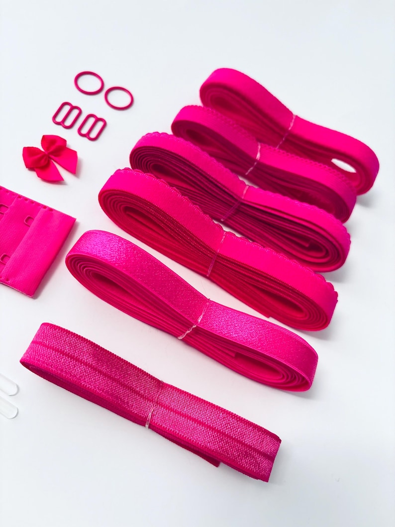 Op de afbeelding: Een set roze bh-bandverlengstukken met een roze strik, twee kleine roze ringen en twee grotere roze gespen. De verlengstukken zijn gemaakt van elastiek en hebben een gegolfde rand.