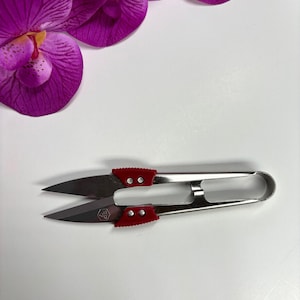 May include: A pair of silver thread snips with red handles, resting on a white surface. The blades are sharp and pointed. A purple orchid flower is in the background.