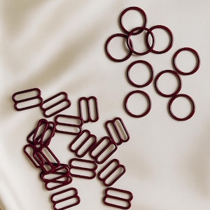 Op de afbeelding: Een verzameling bordeauxrode beha-hardwarecomponenten, waaronder ringen en schuifjes, ligt op een witte satijnen ondergrond. De metalen onderdelen zijn in groepen gerangschikt, waarbij hun vorm en ontwerp worden getoond. De afbeelding benadrukt de details van deze lingerie-onderdelen.