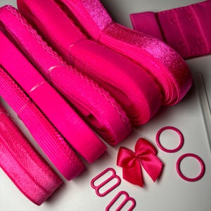 May include: A set of pink bra straps, hooks, and rings. The straps are made of elastic and have a velvet-like texture. The hooks and rings are metal.