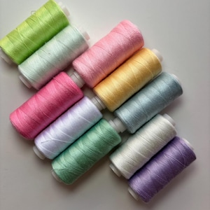 May include: A collection of colorful spools of thread in various pastel shades, including green, pink, yellow, and purple. The threads are neatly wound on white spools, perfect for sewing and crafting projects.