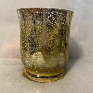 Puede incluir: Un portavelas de vidrio dorado con una superficie agrietada y texturizada. El soporte tiene una parte superior acampanada y una base redonda. El vidrio tiene un acabado metálico reflectante. El portavelas está diseñado para uso decorativo.