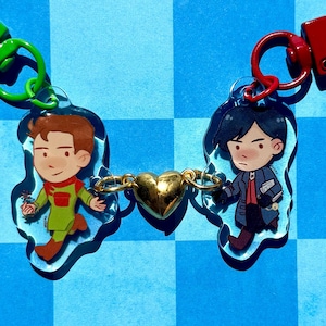 May include: Two acrylic character keychains connected by a gold-colored heart charm. The left keychain features a figure in a green outfit with a lime green clasp, while the right keychain shows a figure in a dark coat with a red clasp.
