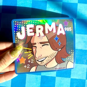 May include: A rectangular holographic sticker featuring a cartoon illustration of a person with brown hair and a beard. The text JERMA 985 is printed in white at the top. The design includes yellow motion lines, an orange star, and the text +2 +2.
