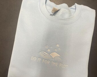Op de afbeelding: Een lichtblauw sweatshirt met de geborduurde tekst "DO IT FOR THE PLOT" onder een ontwerp van een open boek met sterren en cirkels. De sweatshirt is netjes opgevouwen.
