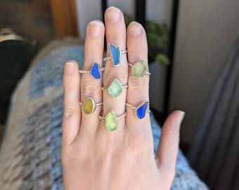 May include: A hand wearing seven rings with different colored sea glass stones. The rings are silver and the stones are blue, green, and yellow.