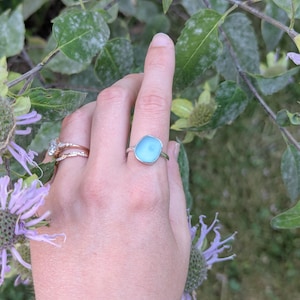 May include: A silver ring with a light blue, irregular-shaped stone. The ring is set against a backdrop of green foliage and purple flowers, creating a natural and elegant aesthetic. The ring is the focal point.