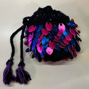 May include: A black crocheted drawstring bag with a purple tassel. The bag is decorated with iridescent pink, blue, and purple scales.