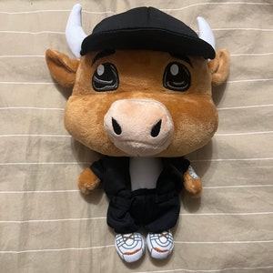 May include: A brown plush bull wearing a black cap, black jacket, and white trainers. The bull has large eyes and a friendly expression.