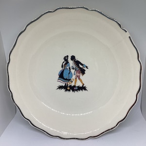 Puede incluir: Un plato de cerámica blanco con un borde plateado presenta una silueta en blanco y negro de una pareja con atuendo del siglo XVIII. La mujer lleva un vestido azul y el hombre lleva una chaqueta marrón y pantalones blancos.