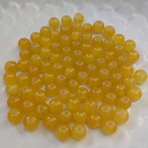 May include: A pile of round, yellow beads made of natural stone. The beads are smooth and polished, and they have a slightly translucent appearance.