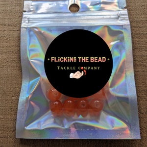May include: A clear plastic bag with a black circle sticker that says "Flicking the Bead Tackle Company" and a red and white hand holding a red bead. Inside the bag are several orange beads.