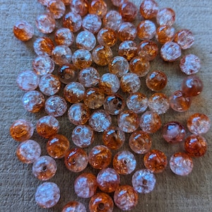 May include: A collection of clear, round beads with a crackled, textured appearance. The beads are a mix of orange and clear, with a hole through the center for stringing. The beads are scattered on a neutral surface.