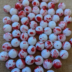 May include: A collection of white round beads with red splatter paint. The beads are approximately 1 inch in diameter.