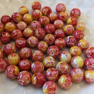 May include: A collection of round beads with a marbled design. The beads are primarily red and yellow, with white veining throughout. Each bead has a hole for stringing. These beads are suitable for jewelry making.