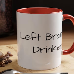 May include: White ceramic mug with a red rim and handle. The mug has black text that reads "Left Brain Drinker".