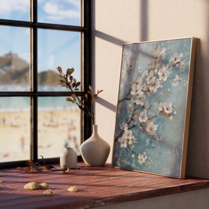 K&ouml;nnte beinhalten: Ein gerahmter Leinwanddruck mit einem wei&szlig;en Blumenarrangement auf blauem Hintergrund. Der Druck lehnt an einer wei&szlig;en Wand neben einem Fenster mit Blick auf einen Strand. Zwei Keramikvasen stehen auf einer Holzoberfl&auml;che vor dem Druck.
