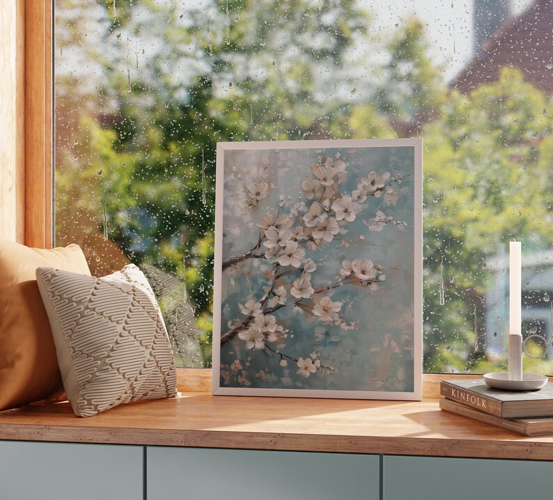 K&ouml;nnte beinhalten: Ein gerahmtes Gem&auml;lde mit einer wei&szlig;en Blumenanordnung auf blauem Hintergrund. Das Gem&auml;lde h&auml;ngt an einer Wand &uuml;ber einem Holzregal mit einem Buch und einer Kerze.