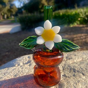May include: A handcrafted glass art piece featuring a flower design. The piece has a brown base with a white and yellow flower and green leaves. The glass is transparent, allowing light to pass through, and is set against a natural outdoor background.