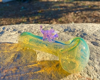 May include: A yellow iridescent glass pipe with a purple glitter dinosaur figurine on top.