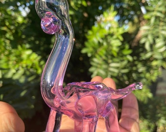 May include: A purple glass dinosaur-shaped pipe with a rounded bowl and a long neck.