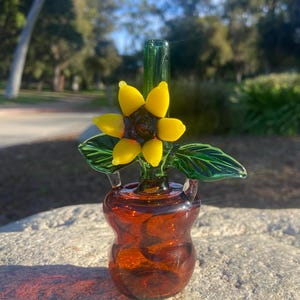 May include: A glass pipe shaped like a sunflower with a brown glass base. The sunflower has green leaves and yellow petals.