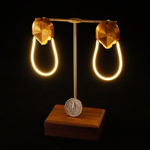 May include: A pair of gold-colored earrings with a light-up feature. The earrings are displayed on a wooden stand with a coin underneath.
