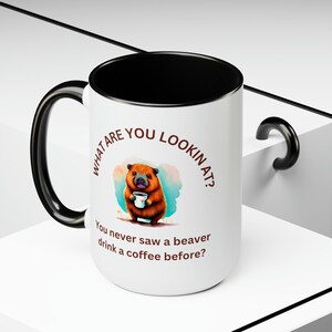 Könnte beinhalten: Eine weiße Keramiktasse mit schwarzem Henkel und Rand. Die Tasse zeigt ein Cartoon-Biber, das eine Tasse Kaffee hält, und den Text "What are you lookin at? You never saw a beaver drink a coffee before?"