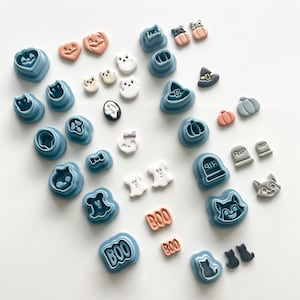 May include: A collection of Halloween-themed cookie cutters and decorative candies. The cutters are blue and shaped like pumpkins, cats, and ghosts. The candies are orange, white, and gray, featuring similar Halloween designs. The word "BOO" is also present.