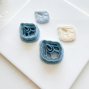 May include: Four polymer clay cutters in the shape of a stylized leaf or petal. Two are light blue, one is off-white, and one is a darker blue. The cutters are arranged on a white surface.