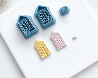 May include: A set of five cookie cutters in the shape of houses. Two of the cutters are blue, one is pink, one is yellow, and one is silver. The cutters are arranged on a white surface.