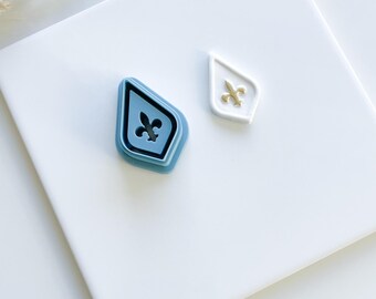 May include: Two blue plastic cookie cutters in the shape of a diamond with a fleur-de-lis design. One cutter is stacked on top of the other.