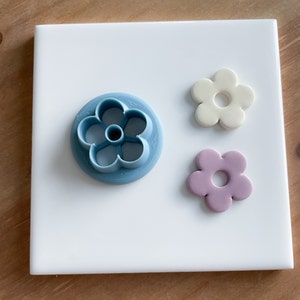 May include: A blue plastic flower-shaped cookie cutter, a white fondant flower, and a purple fondant flower on a white tile surface.