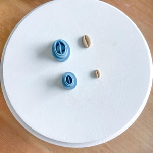May include: Three blue polymer clay coffee bean charms and two brown polymer clay coffee bean charms on a white surface.
