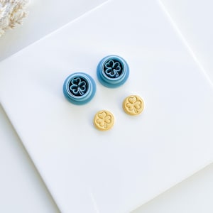 May include: Two blue plastic cookie cutters shaped like four-leaf clovers, and two golden cookies with four-leaf clover imprints.