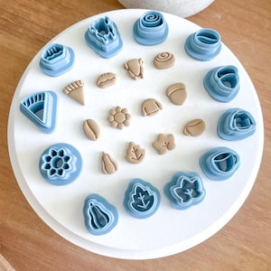 May include: A white circular surface with 20 different cookie cutters in various shapes and sizes. The cutters are made of blue plastic and feature designs such as leaves, pumpkins, acorns, and flowers.