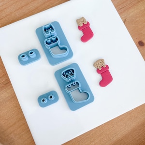 May include: A set of blue plastic cookie cutters shaped like Christmas stockings with dogs peeking out of the top. The cutters are on a white surface. There are two red fondant cookies shaped like stockings with dogs peeking out of the top.