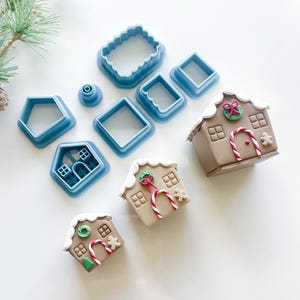 May include: A collection of blue plastic cookie cutters in various shapes, including squares, rectangles, and a house shape. Several decorated gingerbread houses with white icing, candy canes, and festive details are also present.
