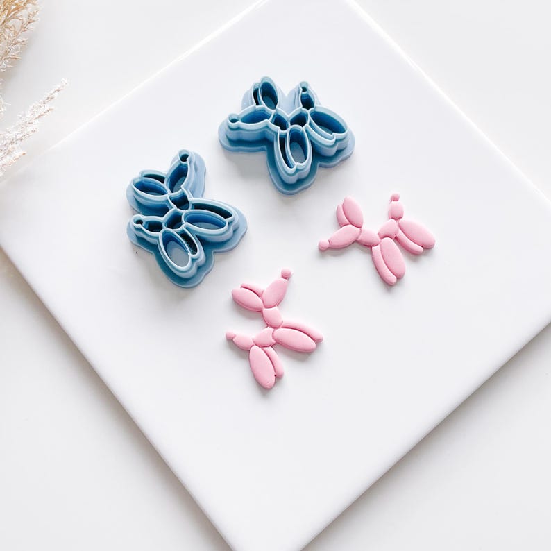 Puede incluir: Cuatro art&iacute;culos con forma de globo. Dos son cortadores de galletas de pl&aacute;stico azul y dos son caramelos rosas con forma de globo. Los cortadores tienen la forma de un grupo de globos. Los caramelos tienen la forma de un solo animal globo.
