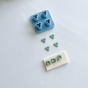 May include: A blue plastic mold with four flower-shaped cutouts. There are five teal-colored flower-shaped clay pieces and three teal-colored flower-shaped clay pieces with gold centers on a white surface.
