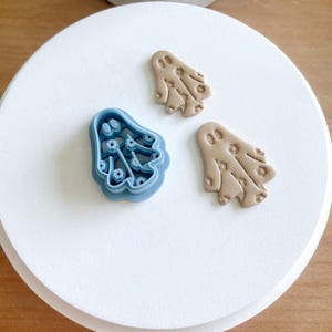 May include: Two ghost-shaped cookie cutters, one blue and one brown, on a white surface. The blue cutter is detailed with a pattern of small circles and the brown cutter has a similar pattern.