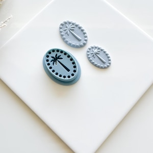 May include: Three blue palm tree-shaped cookie cutters. One cutter is a mold with a palm tree design and a dotted border. Two smaller, solid palm tree-shaped cutters are also present. The cutters are on a white surface.