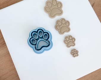May include: A blue paw print cookie cutter next to four tan paw print cutouts of varying sizes. The items are arranged on a white surface. The cookie cutters are likely for making dog or cat-themed treats.